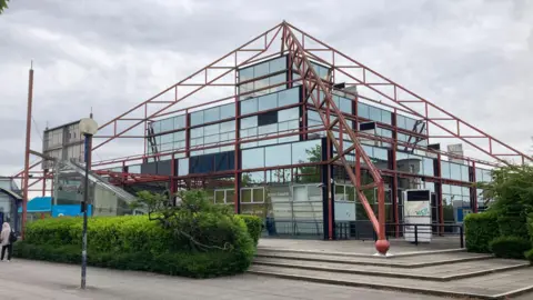 A glass building shaped like a pyramid with red metal support beams lining the edges, this is The Point in Milton Keynes.