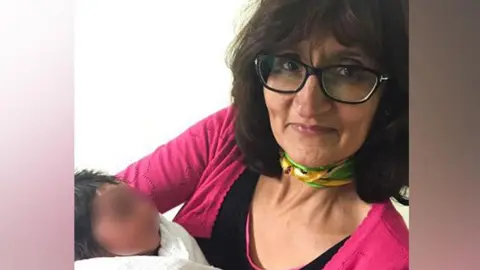 A woman with dark hair, glasses, wearing a pink cardigan holds a small baby in her arms. 