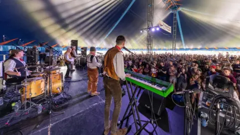 Gorman Photography The Wurzels are shown performing on stage in a large circus style tent, with a large crowd watching on. The photo is taken from behind the band with the crowd looking towards the camera.