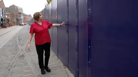 A woman wearing a bright red short-sleeved shirt and black trousers is standing on a cobblestone street next to a tall, solid barrier made of dark blue panels. She is gesturing toward the barrier with one arm extended, as if indicating its length or height. The street has old metal tram lines running along its surface, and older brick buildings are visible in the background on the left side.
