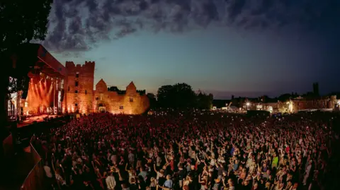 A large crowd of people in the dark with a castle illuminated in the background and a stage on the left hand side