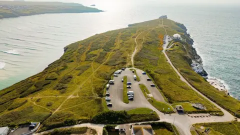 A drone photo taken above East Pentire Headland on a sunny day. The car park is clearly visible in the centre of the peninsula and has about 12 vehicles parked in it. Lewinnick Lodge can be seen further down the headland to the right and there is a house on the water line before the lodge. The headland has scrub and grass with footpaths crossing it. West Pentire headland is visible to the left.