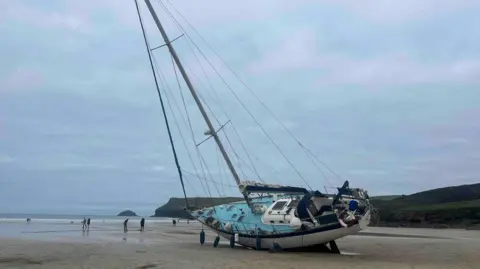 A yacht with a mast and a blue deck on a beach lying on its port side with fenders dangling off the boat and a number of people walking in the background along with a cliff-edge leading to the sea on a cloudy day.