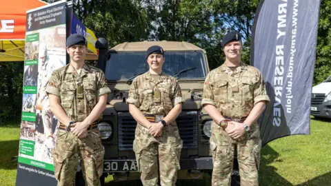 Telford & Wrekin Council Three army personnel stand in front of a military Land Rover in khaki uniform. The two men and a woman are all wearing black berets and standing with their hands clasped in front of them, with banners either side promoting Army Reserves.