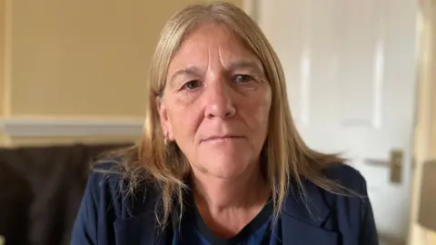 Emma Baugh/BBC Karen Betts who has long blonde hair and is wearing a navy blue jacket over a blue top. She is sitting on a sofa in a sitting room. 