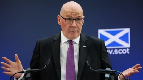 John Swinney, a bald man wearing glasses, a dark jacket, white shirt and purple tie, has his hands outstretched as he speaks into two microphones