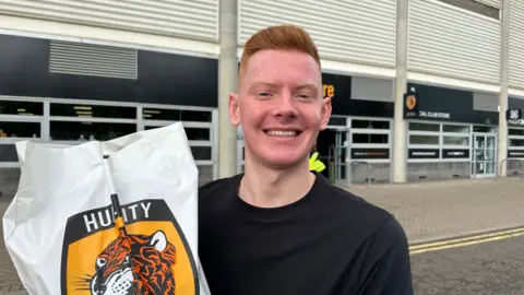 Matt Dean/BBC Joe Collins pictured outside a stadium holding a bag with the club's logo on it. He is wearing a black jumper and smiling at the camera.