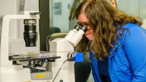 Bowel Cancer UK Dr Osborn, who has long dark hair and is wearing a bright blue shirt,  is looking down a microscope in a lab