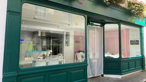 Mill Street Community Café Exterior of the Mill Street Community Café in St Peter Port Guernsey. A dark‑green‑fronted shop with large windows, flower boxes above the entrance, and a view inside showing white tables, chairs and a tiled counter area.