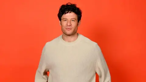 The actor James Norton wearing a white woollen jumper and standing in front of an orange back drop.