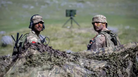 Two soldiers in camouflaged green clothing sitting on some grass. There is netting behind them, in the foreground. They are turned to look at the camera.