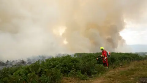 South Wales Fire and Rescue Service Firefighter at work on mountain in yellow helmet and red suit spraying water onto bracken as smoke billows heavily behind him