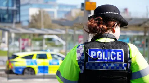 A female police officer in uniform is pictured with her back to the camera. She is wearing a black hat and her red hair is tired into a bun at the back. Beyond her, on the road, is a marked police car. 