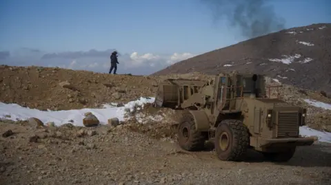 An Israeli soldier stands on top of a rocky hill, some snow seen on the ground, with a dirt-coloured bulldozer below the person 