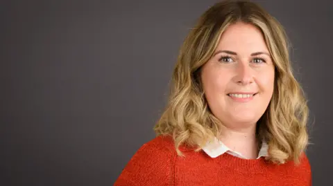 BCP Council Millie Earl in an orange jumper and in front of a neutral background. Her hair is should length, blonde and wavy