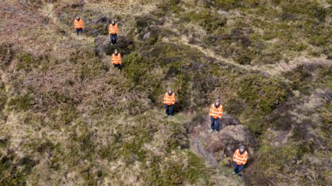 Aerial shot of six people wearing high-vis coats standing in a shallow trench