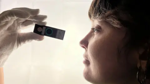 A woman with a nose ring looks at a microscope slide. She has brown hair and is in profile. She is wearing white gloves. 