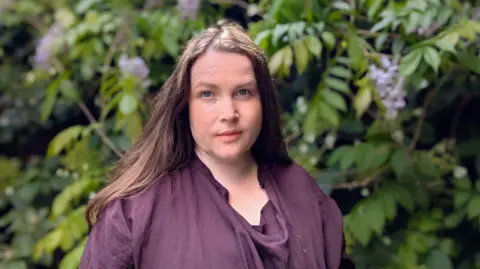 Fiona Mackenzie Woman with brown hair facing the camera with a neutral expression. She is wearing a dark purple top and standing outside in front of a large green bush.
