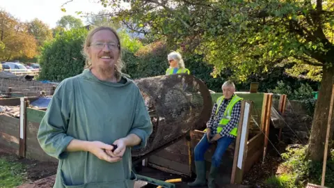 Compost mentor, Ben Bryant, who has auburn, tied back hair and matching beard, wearing a khaki fisherman's cotton tunic with pockets, standing in front of a compost tumbling machine with two volunteers sitting in the background.