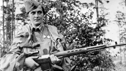 A female soldier, clad in army camouflage, a hat and holding a rifle as she looks away from the camera. The picture is monochrome.