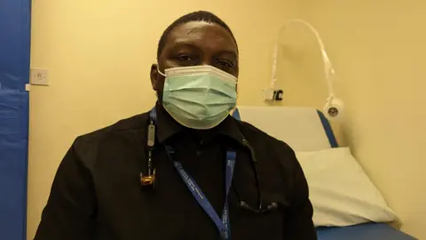 A doctor wearing a black top and a face mask. He has a lanyard and a stethoscope around his neck. He is sitting in a yellow/cream room with a bed behind him.