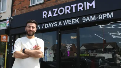 Razwan stands outside his shop. His arms are folded and he is smiling at the camera. His shop frontage in black with a white sign.