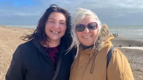 Josh McLaughlin/BBC Lisa Crowther and Ally pose for a photo on Worthing seafront. Blue sky is poking out behind dark grey clouds, as both women smile while their hair blows in the wind.
