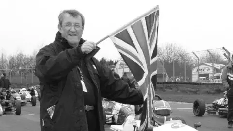 EDP Photo News A black and white image of a man holding up a flag on a race course. Behind him are a number of race cars lining up to start.