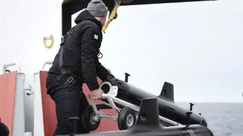 A person in a black work suit holding a black submarine-shaped object on a wheeled trolley on a ship at sea.