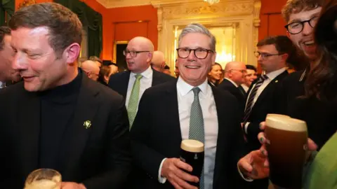 PA Media A man in a navy suit, white shirt and green tie hold a pint glass with black liquid with a cream coloured top.  He has grey hair and glasses. Behind him are dozens of people, out of focus, in an orange painted room.