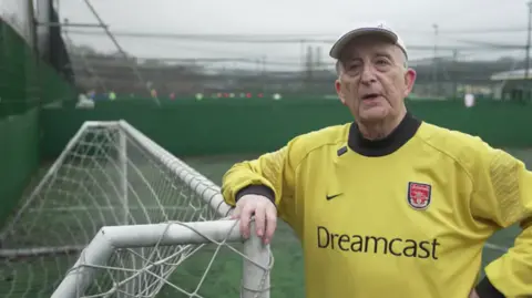 An older man wearing a yellow Dreamcast football shirt leans on a goal. He wears a white baseball can and the pitch can be seen in the background. 