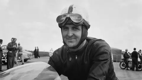 Getty Images Derek Minter smiling while sat on a bike in a black and white image. Other people and old bikes and cars can be seen in the background.