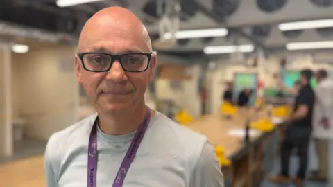 A man wearing a light t-shirt and purple lanyard in a workshop with students working in the distance, behind him.