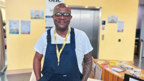 Eastern Education Group Dean stands in a yellow room. He is wearing a white t-shirt under blue overalls. Beside him is a table with books and pens
