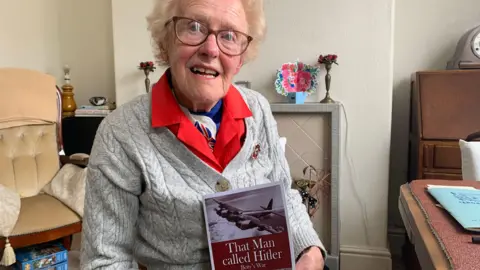 A woman, Betty Quinton, seated in her home. She is slim and wears glasses. She is holding a book entitled That Man Called Hitler.