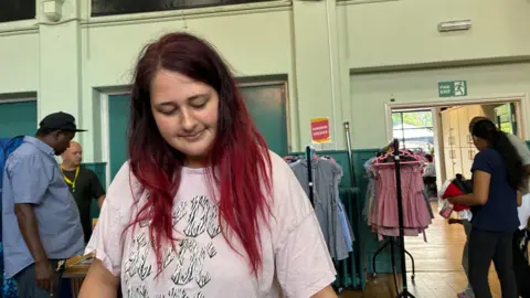 Angelica Jaros has long brown hair that fades to red at the ends. She's wearing a pink ACDC t-shirt and is looking through school uniform with rails of dresses and other people looking at clothes behind her.