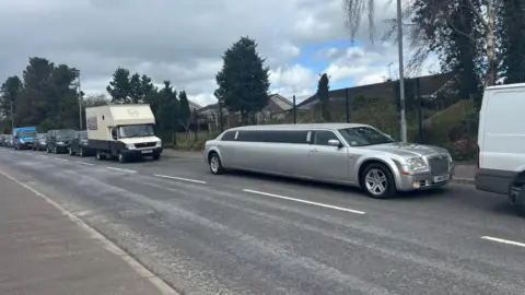 A number of vehicles are sitting bumper to bumber on a road. There is a silver stretch limo, a camper van, some black cars and a blue transit van. 
