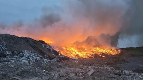Compton Bassett landfill site in the evening, with the burning rubbish a bright orange and huge plumes of smoke. 