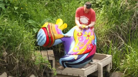 Shaun Dean A man in a red T-shirt holds a large duck sculpture on an angler's wooden platform by a river edge. The duck is painted in rainbow colours with the word 'peace' and coloured flowers along its side.