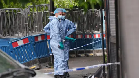 PA Media Two forensic investigators wearing light blue protective suits, face masks, and blue hairnets stand on a pavement behind blue and white police cordon tape. Blue road barriers are visible in the background.