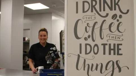 A smiling cafe worker standing behind a table top. She has blond hair which is in a bun and a black chefs coat. There is feature wall that says "I drink the coffee then I do the things"