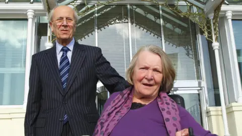 Lord Tebbit stands in a dark blue pinstriped suit with a light blue shirt and striped blue tie in front of a window. His wife Margaret is seated wearing a purple jumper and spotted scarf