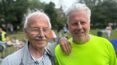 Jordan and Cian's grandfather and dad, John and Glenn. John has white hair and moustache, wearing glasses, grey jacket and striped shirt. Glenn has white hair, wearing a bright yellow t-shirt.