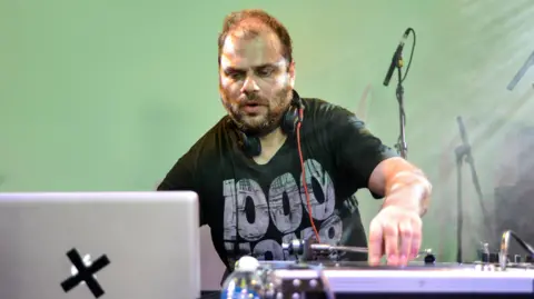 A man DJing behind decks - he has a beard and thin hair 