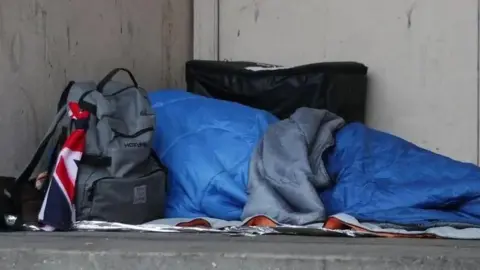 PA Media A person lies on the ground in a blue sleeping bag, rough sleeping. There is a grey backpack next to the person. There is a Union Jack flag tied to the bag.
