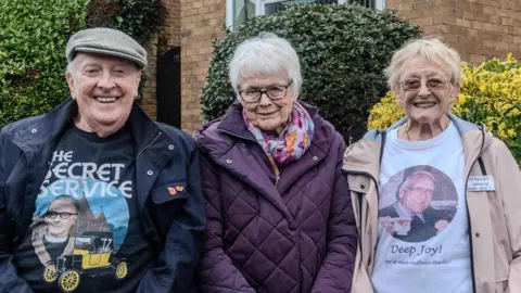 Kate Bradbrook/BBC Lois Johnston is wearing a floral scarf and a purple coat. Either side of her are husband Alex - who is wearing a black T-shirt, navy jacket and flat cap - and sister Marion, who wears a white T-shirt and beige jacket. Stanley Unwin's old bungalow is behind them. It is made of cream bricks and has several carefully trimmed bushes on the front garden.