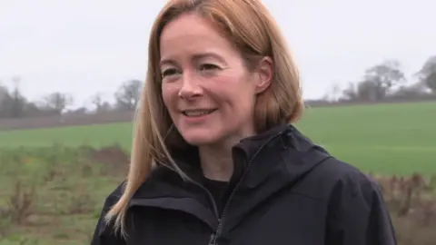 Sarah Mackness, a woman with straight shoulder length hair and a black coat is smiling to the left of the camera. She appears to be standing in a field