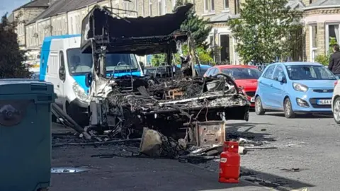 BBC/Andrew Barton The image shows the aftermath of a vehicle fire on a residential street outside a row of terraced houses. The street is lined with parked vehicles. In the centre of the image is a severely burnt-out campervan. The vehicle is destroyed, with the metal frame exposed and blackened. 