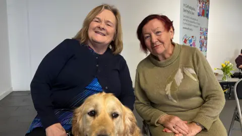 Two older women are sitting next to each other looking at the camera. Judith has blonde hair and is wearing a navy cardigan. In front of her is her golden retriever guide dog. Sitting on the right is a lady with short, red hair and she is wearing a long, green jumper.