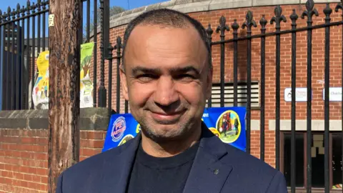 A man with short black hair and short black, greying stubble smiles at the camera. He wears a black T-shirt and navy suit jacket. A school building is behind him.